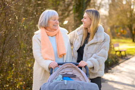 Mother and grandmother smiling at each other while walking with baby stroller in park. Authentic...