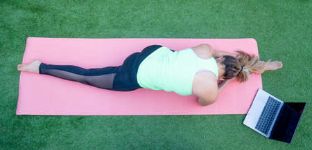 A woman doing an online yoga class in the garden of her house. We can see a close-up of her right leg and arm while the woman performs meditation exercises following the online class on her laptopの写真素材