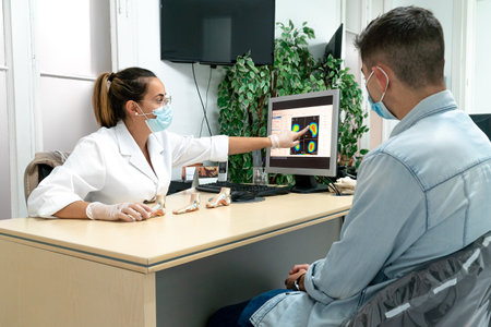 A young podiatrist wearing a mask explains the diagnosis to a young patient in her office. The...