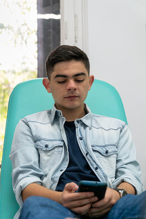 Teenager using his smart phone sitting in a clinic seat while waiting for the doctor to arrive