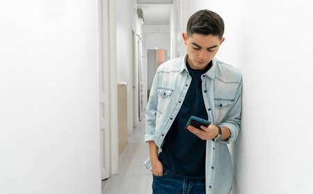 Teenager using a smart phone while waiting at a clinic. He is leaning against the wall with one hand in his pocket and you can see the clinic corridor in the backgroundの写真素材