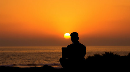 Silhouette of a man with laptop sitting on the beach at sunset.