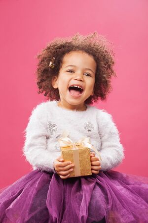 Happy little girl holding a shiny gift box against pink backgroundの写真素材