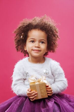 Cute little girl holding a shiny gift box against pink backgroundの写真素材