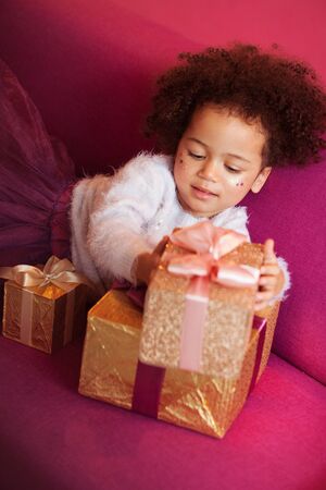 Cute little girl with a lot of gift boxes sitting on sofa against pink backgroundの写真素材
