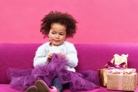 Cute little girl with a lot of gift boxes sitting on sofa against pink backgroundの写真素材