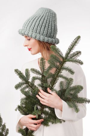 Beautiful woman in warm knit hat holding branch of fir against white backgroundの写真素材