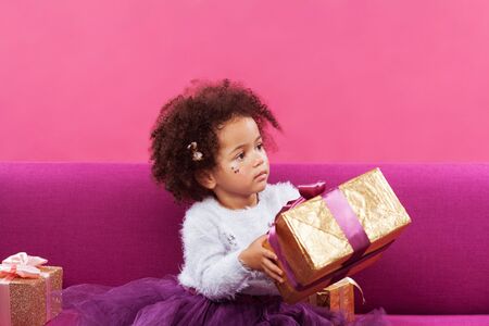 Cute little girl with a lot of gift boxes sitting on sofa against pink backgroundの写真素材