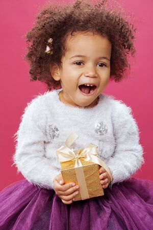 Cute little girl holding a shiny gift box against pink backgroundの写真素材