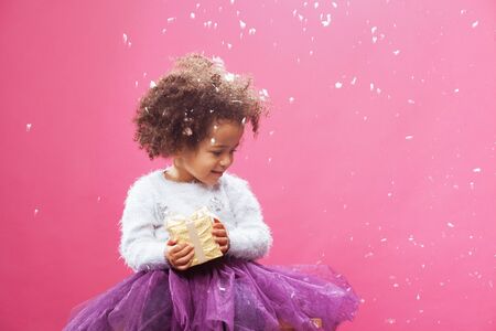 Cute little girl holding a shiny gift box against snowy pink backgroundの写真素材