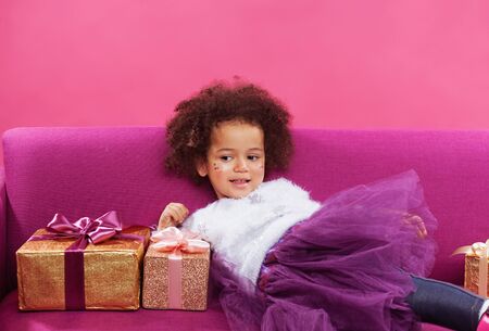 Happy little girl with a lot of gift boxes sitting on sofa against pink backgroundの写真素材