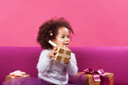 Cute little girl with a lot of gift boxes sitting on sofa against pink backgroundの写真素材