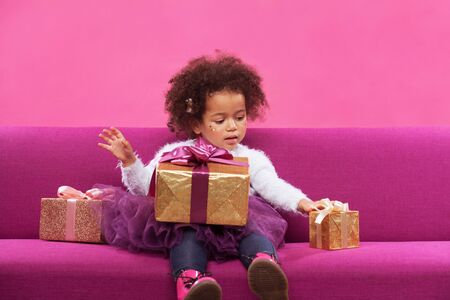 Cute little girl with a lot of gift boxes sitting on sofa against pink backgroundの写真素材