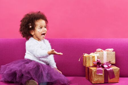 Happy little girl with a lot of gift boxes sitting on sofa against pink backgroundの写真素材