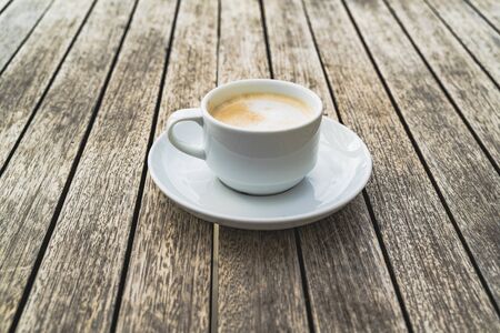 Hot foaming cappuccino in a white ceramich cup on a saucer stands on a table of wooden boards. Coffee is tasty drink and a good measure to feel goodの写真素材