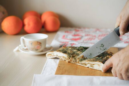 Man slicing up a vegetable pie with big kitchen knife on the cutting board. On the background are orranges and tea cup. Homemade vegetarian food concept. Horizontal and color image.の写真素材