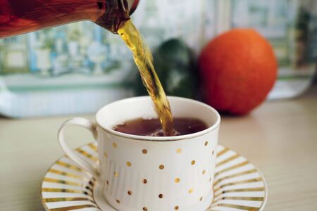 Pouring aromatic tea into a cup from teapot, fruits are on background. Cup and saucer are on wooden table in domestic kitchen. Color image and close up.の写真素材
