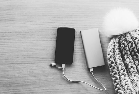 Mobile phone in winter charging from power bank, battery charger, on wooden background. Black and white image, top view. Portable charging device especially helpful while travelingの写真素材