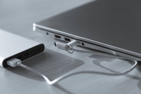 Laptop juicing from battery charger on sunny day, power bank on wood background. Portable charging device very effective while business travel. Black and white image, high angle view and macro. Business still life.の写真素材