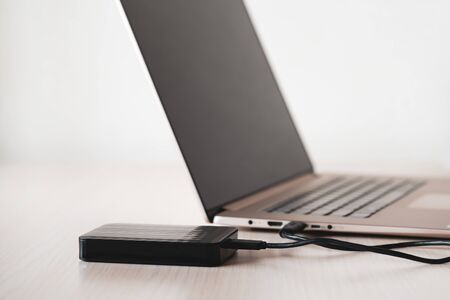 Black external hard disk, hdd on wooden table connected with mini usb cable to laptop, white background. Color image, modern technology concept.の写真素材