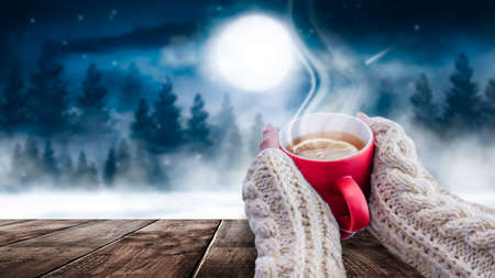 Red cup with coffee, tea in female hands on a snowy background, winter forest. A red cup in hands against the background of a winter forest landscape on soft, knitted sleeves. Coffee, tea on a woodenの写真素材