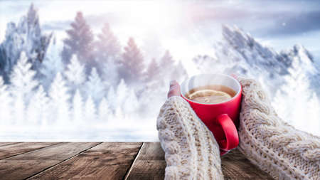 Red cup with coffee, tea in female hands on a snowy background, winter forest. A red cup in hands against the background of a winter forest landscape on soft, knitted sleeves. Coffee, tea on a woodenの写真素材