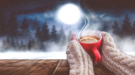 Red cup with coffee, tea in female hands on a snowy background, winter forest. A red cup in hands against the background of a winter forest landscape on soft, knitted sleeves. Coffee, tea on a woodenの写真素材