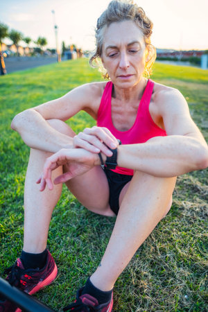 A woman in a red tank top is sitting on the grass and looking at her watch. She is focused on the time and possibly waiting for someone or somethingの写真素材