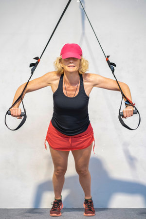A woman is exercising with a trx resistance band. She is wearing a black tank top and red shorts