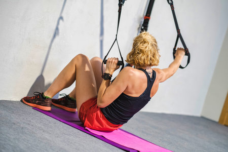 A woman is exercising with a resistance trx band. She is wearing a black tank top and red shorts....
