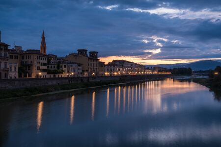 Florence old historical buildings reflecting in Arno river during a overcast sunrise morning with warm sun light with dark cloudsの写真素材