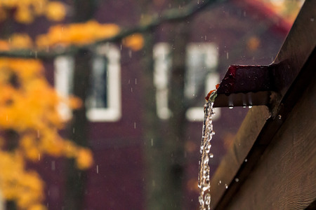 Rainwater pouring from rooftop spout with cedarwood house and autumn leaves blurred in the background.の写真素材