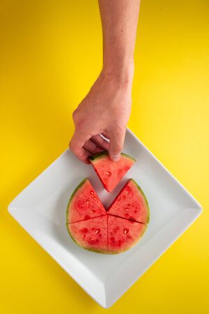 Hand reaching for sliced watermelon on a plate against yellow background.