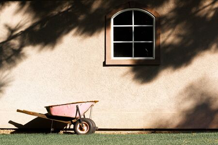 Pink wheelbarrow against a wall with a window and grass.の写真素材