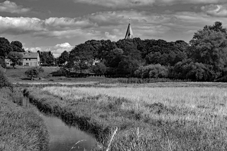 A brook runs through a field. In the background are trees and buildings.の写真素材