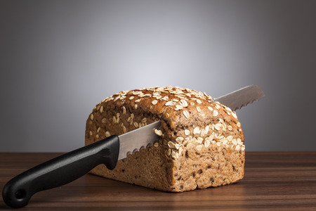 Loaf of wholemeal bread with cutting knife on table in front of gray background.