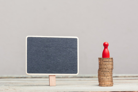 a square blackboard sign (empty, copyspace) next to a stack of eurocents with red pawn on a wood table. grey background, studioshot.の写真素材