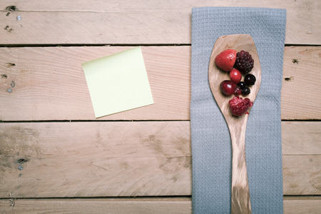 Red and blue berries on cooking spoon and yellow note with copy space, glass of milk, high angle birds eye view on wood table planks.の写真素材