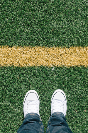 Artificial turf grass with yellow side line on sports field with two shoes, personal perspective from above, footsie or flortraitの写真素材