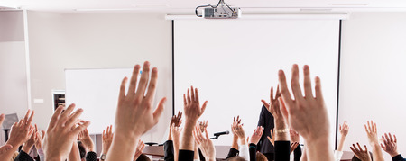 Raised hands and arms of large group of people in class room, audience voting in professional...