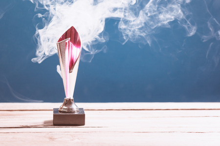 Sports trophy cup on wood desk with dramatic strong contrast light and smoke on blue background,...