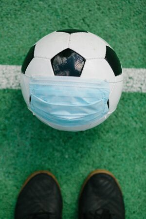 Soccer football with surgical face mask on middle line of artifiical turf field, ready for kick off, symbol image for sports during the corona covid-19 pandemic in 2020の写真素材