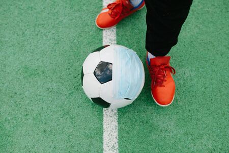 Feet and shoes of boy kicking off soccer football with surgical face mask on artifiical turf field, symbol image for sports during the corona covid-19 pandemic in 2020の写真素材