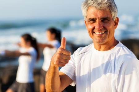 active mature man giving thumb up on beach with family exercising on backgroundの写真素材