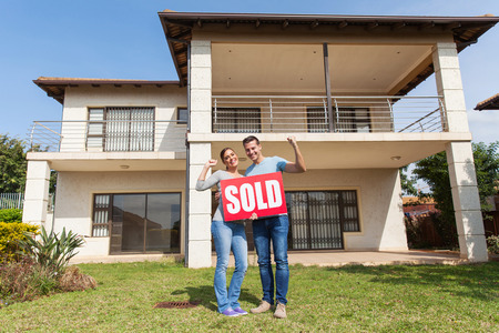 happy couple standing outside their house and holding sold signの写真素材