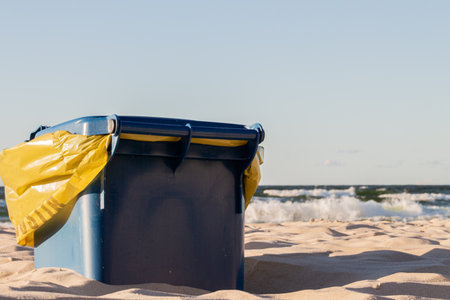Blue beach waste bin with a yellow bag on sandy shore during a bright day near the ocean waves