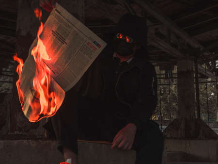 A guy in a mask and coat holds a burning news newspaper, sitting in the catacombsのeditorial素材