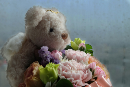 Bear angel doll holding flowers with background of blurry raindrops on the glass window in raining seasonの写真素材