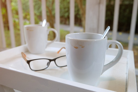 Stained used white glass of coffee on square table with a eye glasses at soft morning lightの写真素材