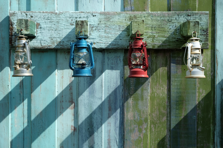 Many storm lanterns with different colors hanging on the old wood blue and green fence wall with the shadow from the sunlightの写真素材